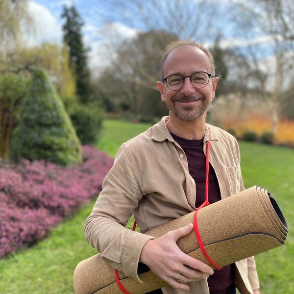 A smiling man holding a rolled yoga mat in a garden with greenery and flowers.