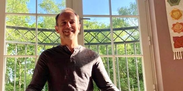 A man smiling sitting in front of large windows with greenery outside.