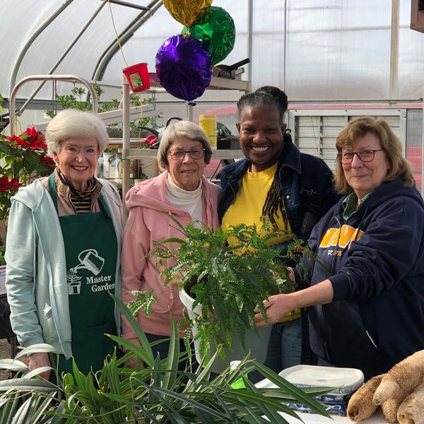 Four women smiling with plants in a greenhouse setting.