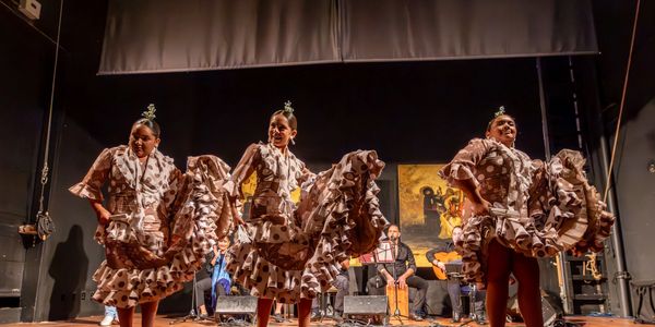 Three women in traditional flamenco dresses performing on stage with musicians behind them.