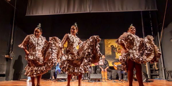 Three women in traditional flamenco dresses performing on stage with musicians behind them.