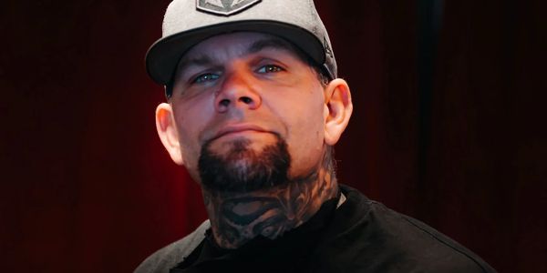 Man with tattoos and a cap poses confidently against a dark background.