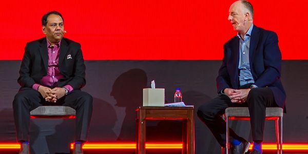 Two men in formal attire sitting on stage with red background.