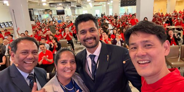 Four smiling people take a selfie in front of a large audience dressed in red.