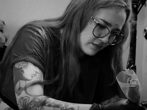 Woman with glasses focused on tattooing, showing detailed tattoos on her arm.