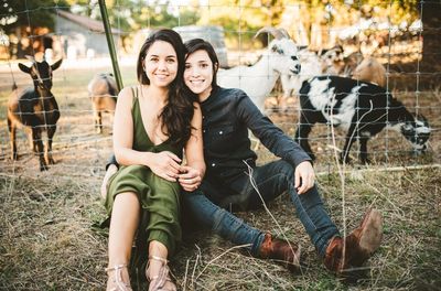 Two women sit in a field with goats behind them.