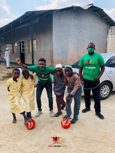 Two men and four boys pose with soccer balls outside a building.