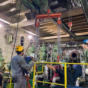 Workers operate heavy machinery with overhead chains in an industrial setting.