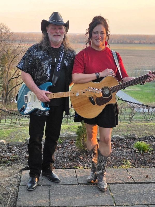 Two musicians with guitars posing outdoors in casual country style.