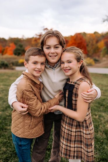 Three children hugging outdoors with autumn trees in the background.