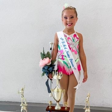 Young girl in a gymnastics outfit with trophies and flowers.