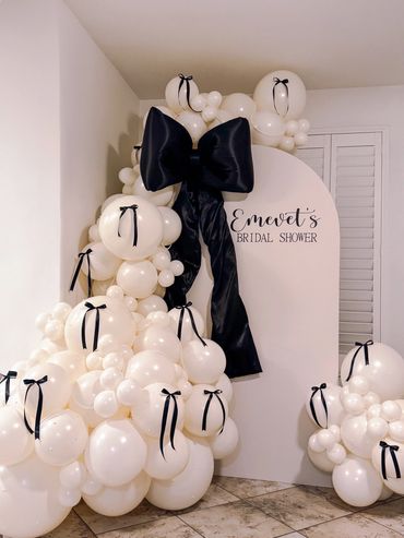 Ivory organic balloon garland with pearl accents and black bows on a wood Chiara backdrop. 