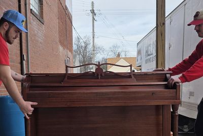 Two men moving a wooden piano on a dolly outside a building.