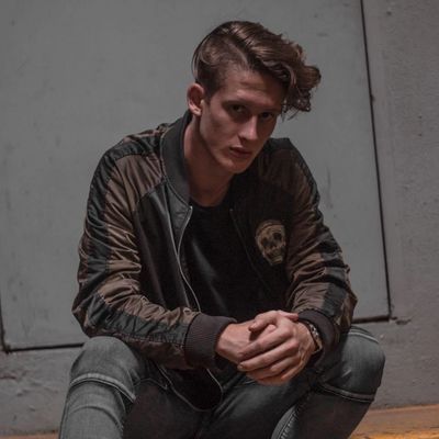 Young man in a bomber jacket sitting with hands clasped, looking seriously at the camera.