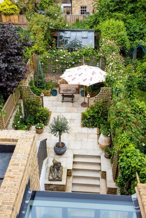 Charming garden patio with umbrella, seating, and lush greenery surrounding a paved area.