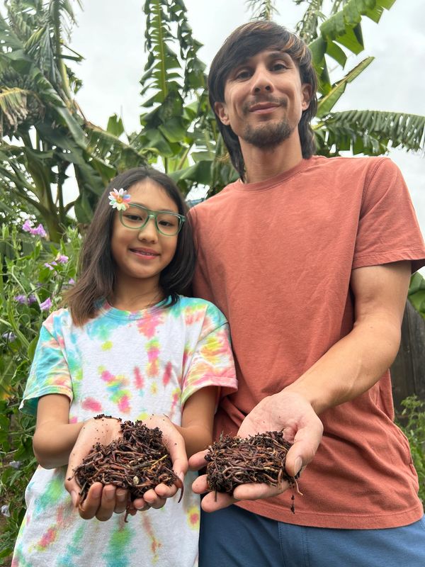 A man and a girl smiling outdoors with plants in the background.