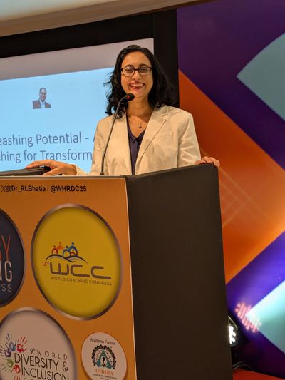 A woman speaker at the World Coaching Congress podium, smiling confidently.