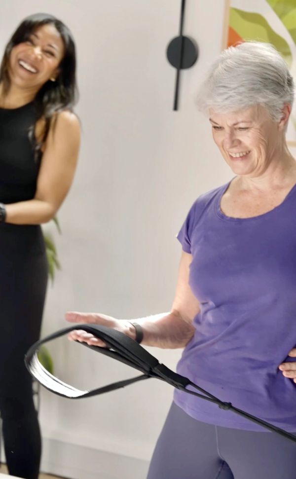 Two women smiling, one holding a strap during exercise.