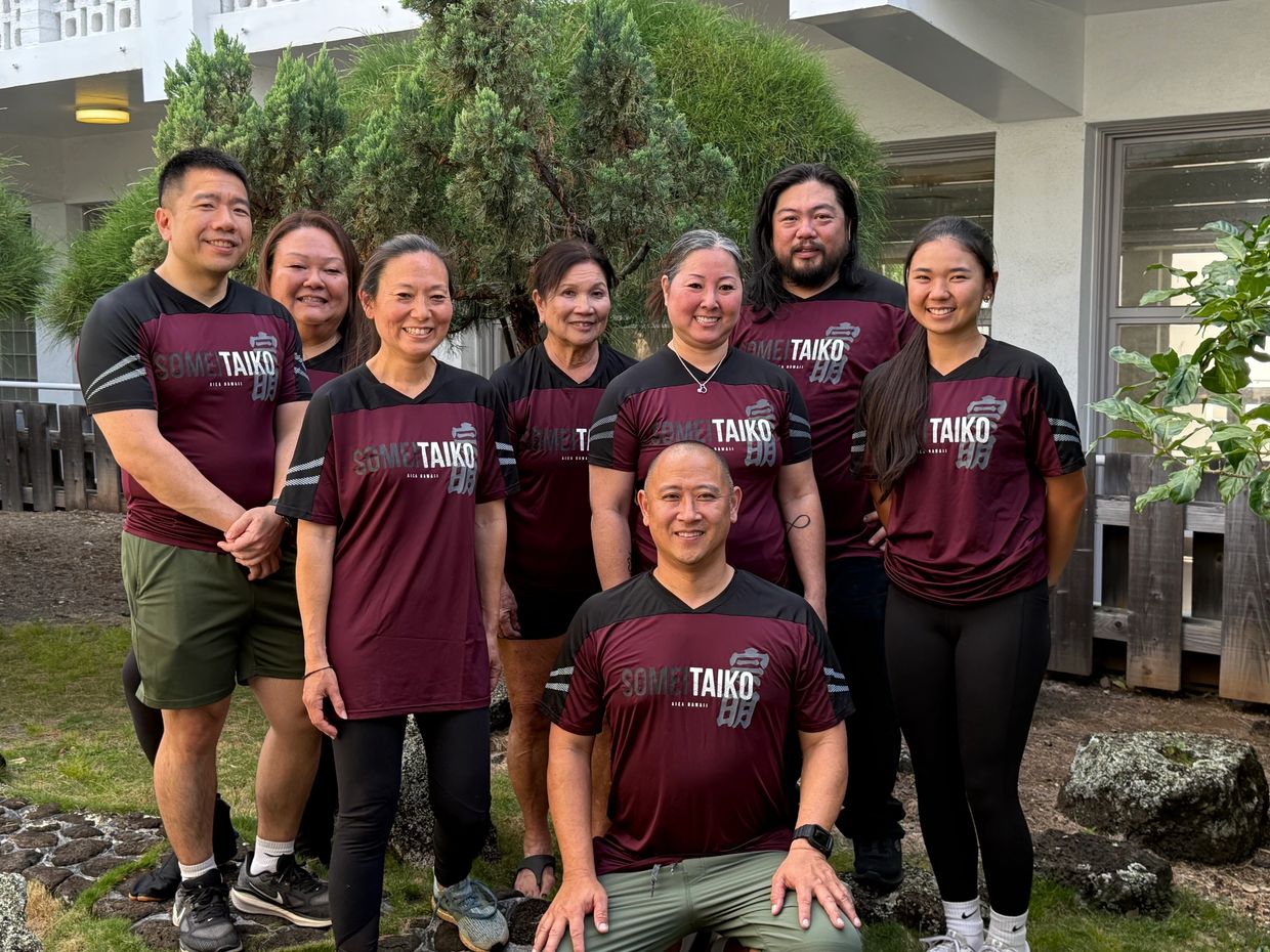Group photo of nine people wearing matching maroon and black TAIKO shirts outdoors.