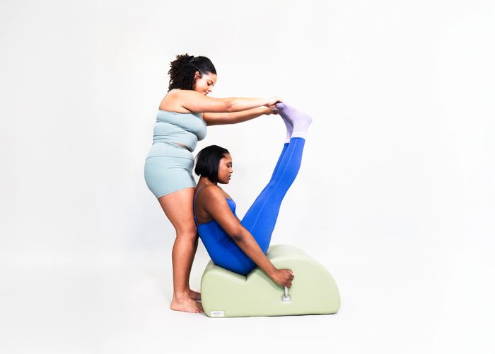 Two women practicing assisted Pilates using a green exercise prop against a white background.