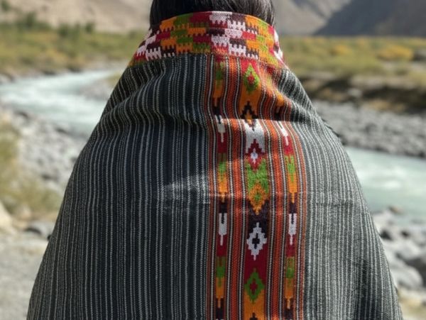 Person wrapped in a colorful, patterned shawl by a river with mountains in the background.