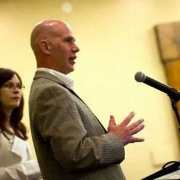 Man speaking at a microphone with a woman in the background.