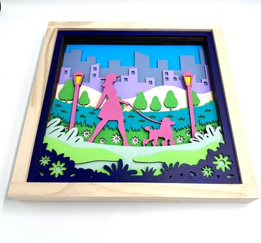 Layered wooden artwork of a woman walking a dog in a park with city skyline.