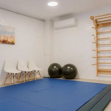 Minimalist exercise room with blue mats, white chairs, and wooden gymnastic rings on a ladder.