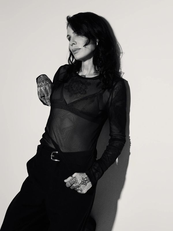 Black and white portrait of a tattooed woman in sheer top and dark pants.