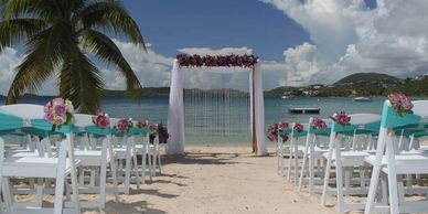 Beach wedding setup with decorated chairs and floral arch by the ocean.