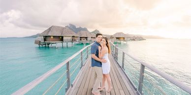 A happy couple embraces on a wooden pier over turquoise water with overwater bungalows in the background.