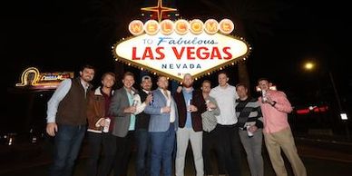A group of men posing in front of the Welcome to Fabulous Las Vegas sign at night.