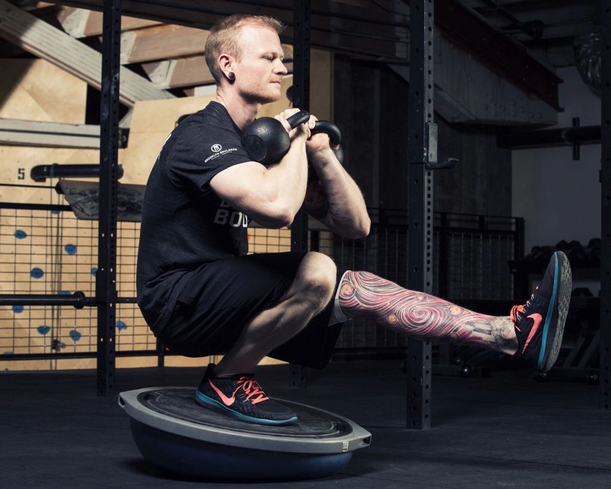 Matt balances in a pistol squat on a BOSU ball while holding two kettlebells at his chest