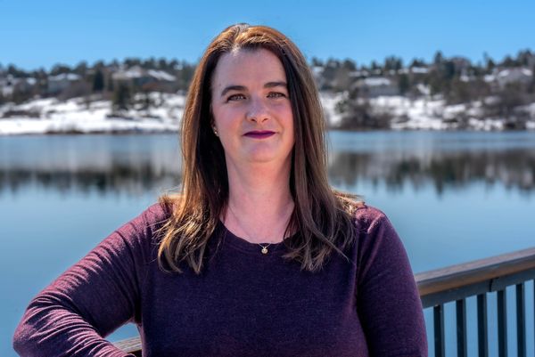 White middle aged female with long brown hair standing in front of a lake with snow on houses and tr