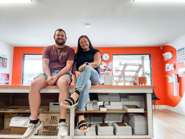 A smiling man and woman sitting on a table in a bright, organized room.