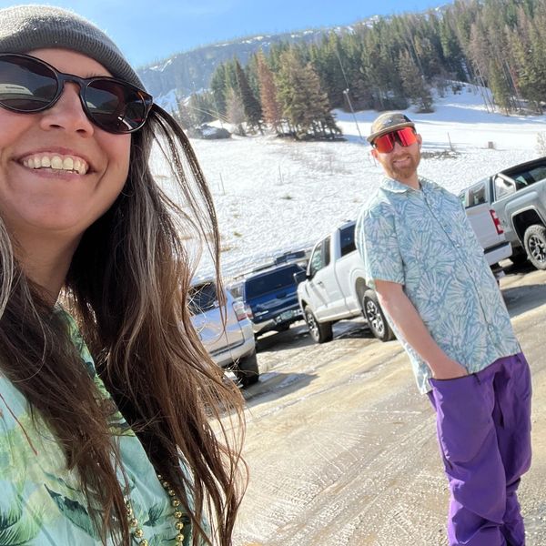Two people in colorful outfits enjoying a sunny day in a snowy mountain parking lot.