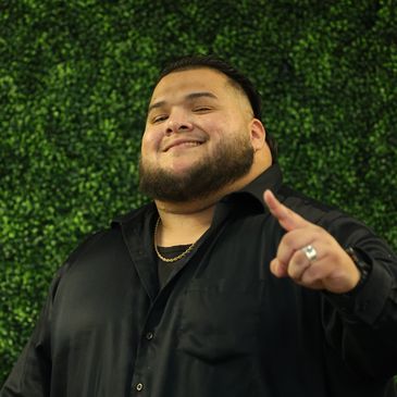 Smiling man in black shirt giving thumbs up with green leafy background.