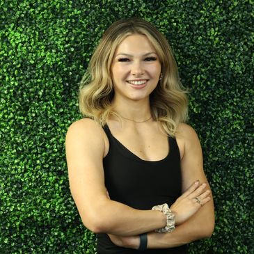 Smiling woman with blonde hair in black tank top standing confidently against green leafy background.