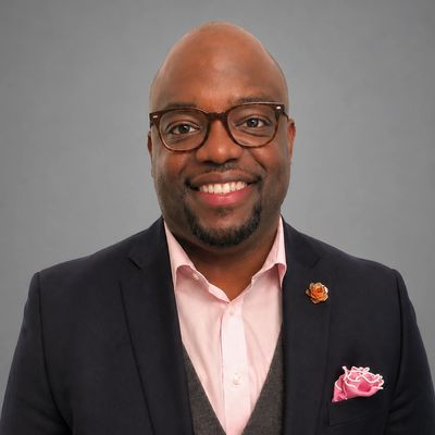 Smiling man in glasses wearing a suit with a pink pocket square and lapel pin.