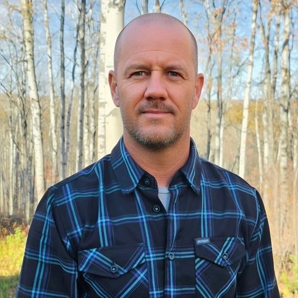 Man in a blue plaid shirt standing in the woods.