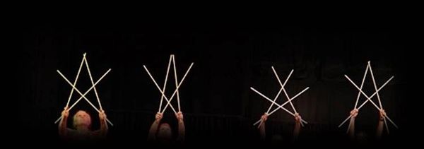 Six hands holding drumsticks forming abstract shapes against a black background.