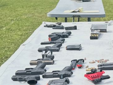 Several handguns and ammunition laid out on tables outdoors.
