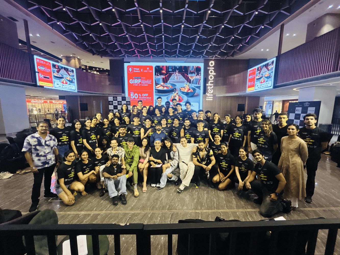 Large group of people posing indoors in front of digital screens and a modern ceiling.