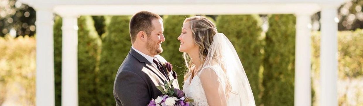 A newly married couple, the bride holding a purple bouquet, smiling at each other outdoors.
