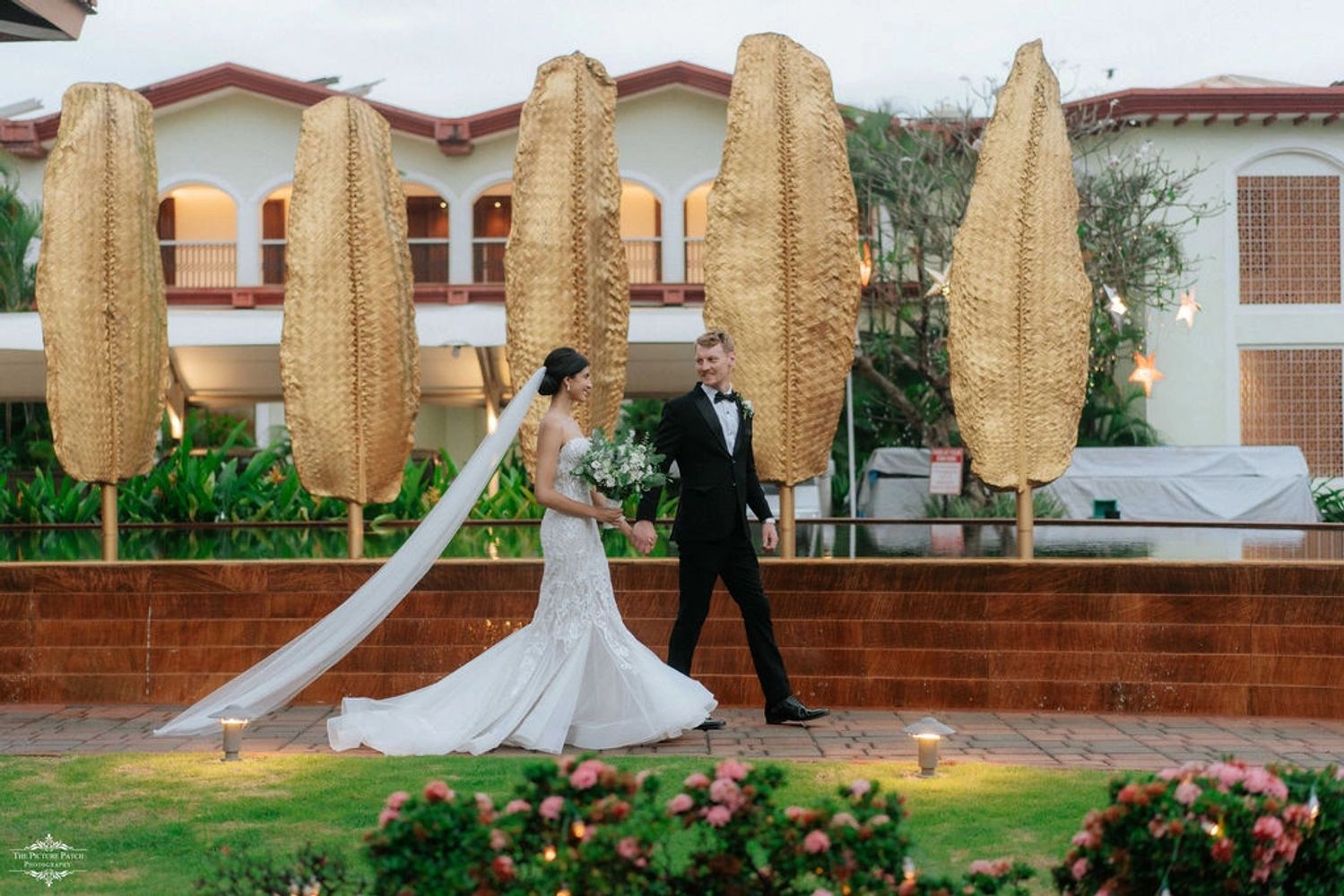 Bride and groom walk hand in hand outdoors with golden leaf sculptures in the background.