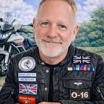 Smiling man in a military-themed jacket with patches and a motorcycle in the background.