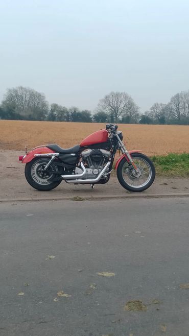 Red motorcycle parked on a rural roadside with fields and trees in the background.
