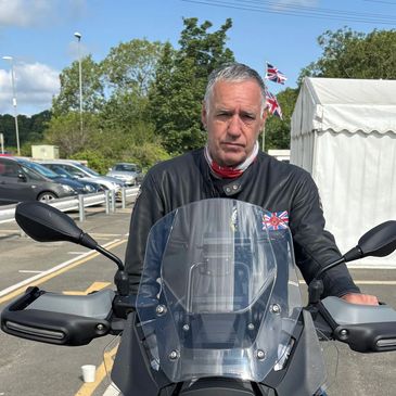 Man in leather jacket sitting on a motorcycle with British flags in the background.