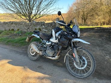 A rugged adventure motorcycle parked on a sunlit country road beside a stone wall.