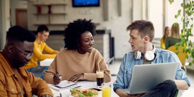 Two students discussing work with laptops and notes in a casual workspace.